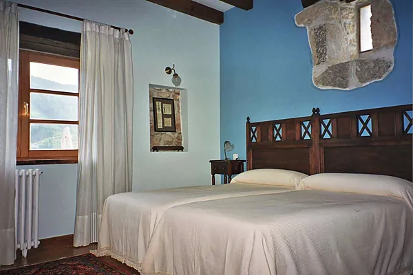Habitación de paredes azules con cabecero de madera tallada, ventana con marco de piedra y cortina blanca.