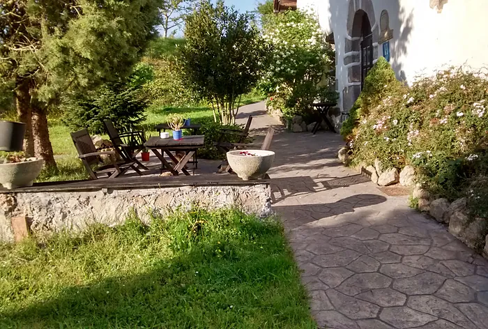 El camino empedrado del jardín, con bancos de madera bajo los árboles y la casa a la derecha.