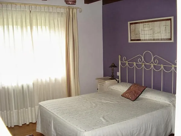 Habitación de paredes lilas con cama de forja blanca y cortinas claras.